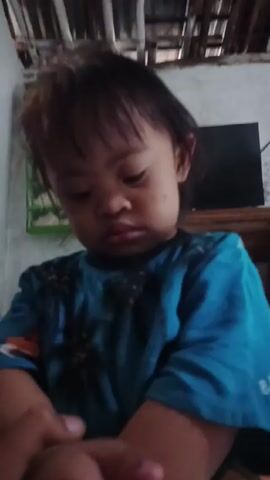A young child in a blue patterned shirt looks down at her hands, then looks up at the camera and speaks. She moves closer to the camera, showing her face and mouth more clearly. The background includes a white wall, a partially visible shelf with items on it, and a glimpse of a ceiling made of what appears to be natural materials like bamboo.