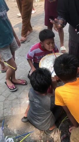 The video shows a group of children gathered around a large, shallow bowl filled with a white substance and thin, red cylindrical objects. One child, wearing a pink and white striped shirt, is kneeling and actively using a straw to move one of the red objects around in the bowl. Another child in a gray shirt is sitting behind the bowl, and a third child in a yellow shirt is observing. Several other children and adults are visible in the background, standing and watching the activity. The ground appears to be paved with bricks, and there are scattered debris, including chalk or white powder, on the ground. The scene takes place outdoors in what seems to be a public area or courtyard.