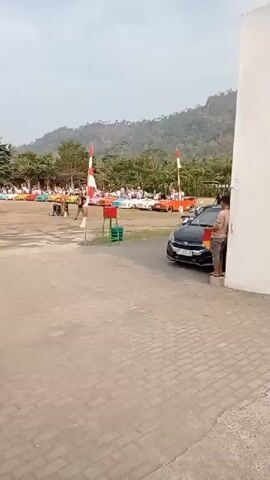 The video shows an outdoor event with numerous colorful small cars lined up in the background, resembling a car show or parade. A group of people are visible standing around the cars and in the middle ground. Two tall poles with red and white striped flags are positioned in the foreground. A white building is on the right side of the frame, and a black car is parked near it. In the immediate foreground, a person in a dark shirt and pants is standing. The ground is paved with bricks and concrete. Hills and trees form the backdrop under a pale sky.