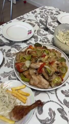 The video displays a table laden with food, including plates of roasted chicken, french fries, and a salad composed of tomatoes, cucumbers, and onions. There's also a bowl of shredded cabbage salad, bread, crackers, and a bowl filled with wrapped candies. The scene appears to be a mealtime gathering. A person's hand is visible in the frame, using a fork to pick up a piece of chicken from a plate. The table is covered with a patterned tablecloth.