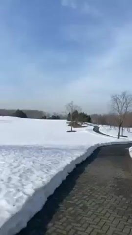 The camera pans across a snowy landscape on a bright, sunny day. The ground is covered with snow, with a few patches of green grass visible in the distance. There are trees scattered throughout the scene, and a paved path runs through the middle of the frame. The sky is blue with a few wispy clouds.