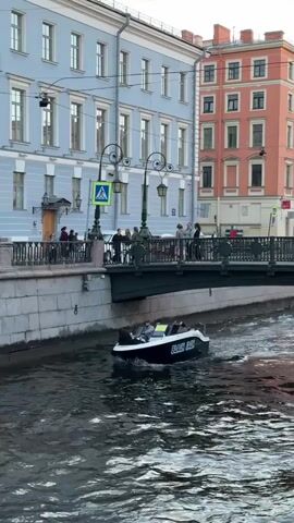 A black and white speedboat with the name "BLACK JACK" visible on its side travels down a canal. The boat is carrying at least five passengers. The canal is bordered by a stone embankment with a decorative railing on top. Behind the embankment, a multi-story building with light blue and orange facades can be seen. A pedestrian bridge crosses the canal in the background, with several people standing on it. The water in the canal appears choppy, with visible wakes trailing behind the speedboat. The sky above is overcast.