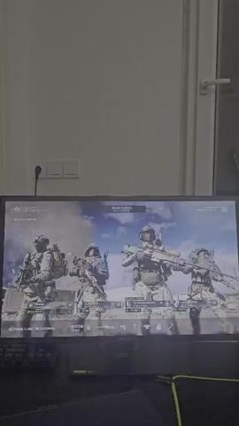 The video captures a computer monitor displaying a video game's pre-game lobby screen. Four characters, depicted as soldiers in tactical gear, are prominently featured. The environment on the screen appears to be an outdoor setting with debris and smoke, suggesting a battlefield or warzone theme. The monitor is placed on a desk, with a portion of a keyboard visible in the foreground. The lighting in the room is subdued, creating a slight glare on the screen, indicating the video was likely recorded in low-light conditions. The background of the room is out of focus but suggests a typical domestic or office setting with a white wall and a door frame. The video is static, with no movement within the frame other than the subtle flicker of the monitor display.