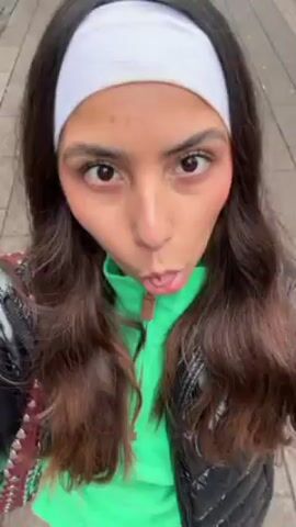 A young woman is recording a selfie video as she walks down a paved street. She wears a white headband, a bright green top, and a shiny black puffer jacket. A burgundy studded bag is slung over her shoulder. Buildings and other pedestrians are visible in the background. The camera angle shifts slightly as she moves.