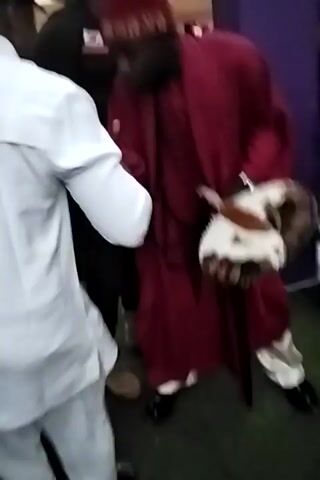 A close-up, blurry video shows a man in a maroon robe and patterned headpiece lying on a green surface. Other people are visible, including one in a purple dress seated on a purple surface and one in a white shirt. Someone touches the man in the robe. Various personal belongings are scattered around the area. The focus shifts erratically, making it difficult to discern details.