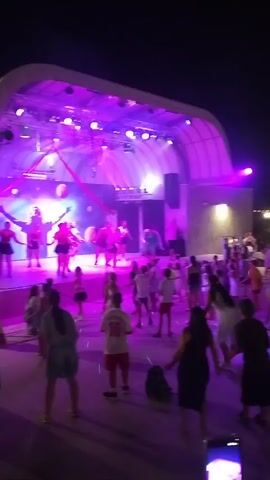 A nighttime outdoor event is captured in the video. A stage with purple and green lighting is visible on the left side of the frame. Several individuals are performing on the stage. A large crowd of people, including adults and children, are gathered in front of the stage, dancing and enjoying themselves. String lights are strung across the background, illuminating the area. The overall atmosphere appears festive and lively.