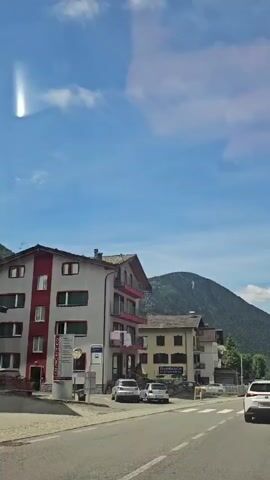This is a first-person perspective video from a vehicle traveling down a paved road in a town. On the left side of the road, there are multi-story buildings with balconies and parked cars. A bus stop sign is visible. On the right side, more multi-story buildings line the road, some with stone facades. In the background, a large, tree-covered mountain rises under a bright blue sky with scattered clouds. The vehicle is following a white SUV. The road has lane markings and a crosswalk. The video appears to be filmed during the daytime.