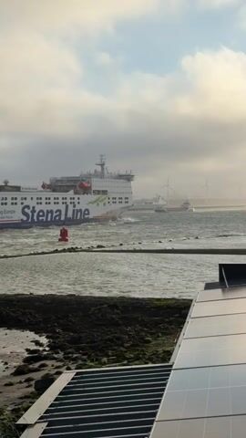 A large ferry named Stena Line is traveling through choppy water near a shoreline. The ferry is white with red and green accents and displays the text "Connecting Europe" and "www.stenaline.com". A red buoy is visible in the water in front of the ferry. In the foreground, a section of a roof covered in solar panels is visible. In the background, wind turbines can be seen on the horizon. The sky is overcast with some visible clouds.