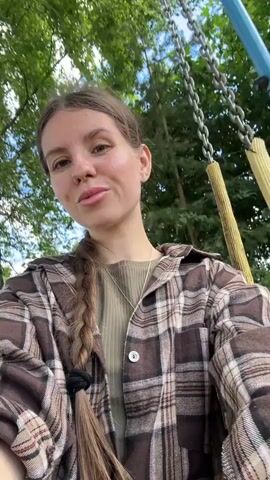 A young woman with long brown hair in a braid, wearing a plaid shirt over a green t-shirt, sits on a swing and records herself with a phone. She is outdoors in a grassy area with trees in the background. The swing set has yellow and blue poles and metal chains. The woman smiles and looks at the camera as the swing moves.