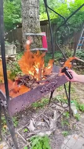 The video captures an outdoor scene in what appears to be a backyard or garden area. A person's hand is visible on the right side of the frame, holding a black device that resembles a torch lighter. This device is being used to ignite a fire in a rusty metal barbecue grill. The grill is filled with wood and some unidentifiable items, which are engulfed in bright orange flames and producing a significant amount of smoke.

Behind the grill, a large tree trunk is prominent. Attached to the tree, at a higher level than the grill, are two items wrapped in what looks like red plastic or fabric, secured with wire. The surrounding area is overgrown with green foliage and leaves, suggesting a natural and somewhat unkempt setting. In the background, a wooden fence and more trees are visible, hinting at a rural or suburban location. A plastic bottle, possibly containing liquid, is placed on the ground to the right of the grill.

The activity is the lighting of a barbecue fire. The flames are intense, and the smoke billows upwards. The presence of the torch lighter and the act of starting a fire suggest a preparation for cooking or a demonstration of fire-starting. The overall mood is dynamic due to the active flames and smoke. The scene is well-lit, implying it is daytime, and the presence of green leaves suggests warm weather, possibly spring or summer. The location context provided is "Правдист-1 Снт, Russia," which is a garden cooperative in Russia. No visible text is present in the video.