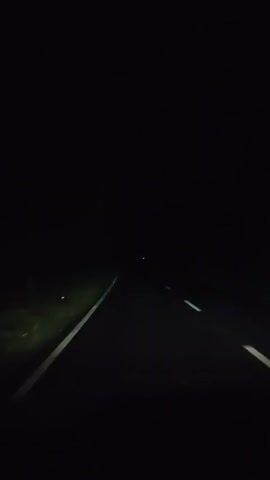 The media file captures a view from the perspective of a moving vehicle at night. The primary subject is a dark, paved road with white dashed lines indicating lanes. To the left of the road, there is a grassy embankment illuminated by faint green light. The sky is completely black, suggesting it is nighttime, and the weather appears clear as there is no visible precipitation or fog. The scene is otherwise devoid of any distinct objects or people, creating a sense of isolation and a focus on the journey.

The activity taking place is travel along this road. The dashed white lines are receding into the distance, and a single, faint light is visible far ahead, hinting at a distant destination or another vehicle. There are no visible signs of human presence apart from the implied driver and passenger of the vehicle from which the recording is made. The overall mood conveyed is one of quiet movement through darkness, perhaps evoking feelings of anticipation or solitude. The location context, Teupinreusep, Indonesia, does not offer any specific visual cues within the frame itself, as the scene is dominated by the dark road and the sparse illumination. No text is visible in the media file.