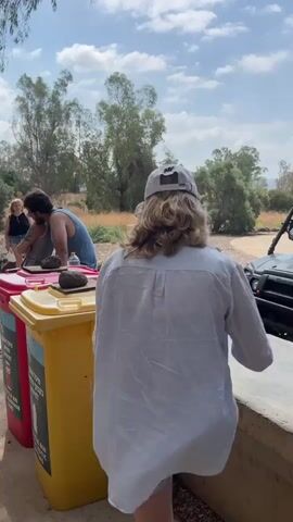 The video shows an outdoor scene with several people. In the foreground, there are two large recycling bins, one red and one yellow. On top of the red bin, a man in a blue tank top is leaning over and appears to be interacting with a person sitting on the bin's edge, who is wearing a grey tank top and denim shorts. In the background, there are more people standing and sitting, and a wooded area with trees and dry grass. The sky is partly cloudy. The overall impression is of a casual gathering or event in an outdoor setting.