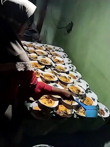 A person wearing a dark headscarf and a red shirt is serving food from a blue ladle onto numerous plates arranged in a pyramid on a table. The plates are white with a decorative pattern and contain a yellowish curry-like dish. The environment appears to be indoors with green walls and dim lighting.