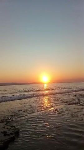 The video shows a sunset over the ocean. The sun is a bright orange orb low on the horizon, casting a shimmering reflection on the water. Gentle waves lap against a rocky shore in the foreground. The sky transitions from a warm orange near the sun to a pale blue at the top. There are no people or objects visible in the video besides the natural elements of the beach and the ocean.