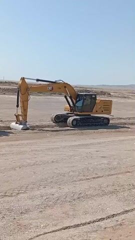A yellow CAT excavator is digging a trench in a dirt field under a clear blue sky. A person wearing a hat and light-colored clothing is walking in the background, away from the excavator. The ground is mostly dirt and gravel.