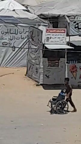 A young boy pushes a wheelchair occupied by a person, moving from left to right across the frame. The scene appears to be an outdoor market or settlement, with tents and makeshift structures in the background. The ground is dusty and sandy.