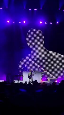 A male singer stands on a stage with a guitar, bathed in purple light. Behind him, a large screen displays a pixelated, black and white image of his face. The screen is divided into a grid of small, white squares, creating a mosaic effect. The singer is wearing a white shirt and white pants. Two other figures are visible on stage behind him, one playing a keyboard. A crowd of people is visible in the foreground, their faces illuminated by the stage lights. Some audience members are holding up their phones to record the performance. The overall atmosphere is that of a live concert.
