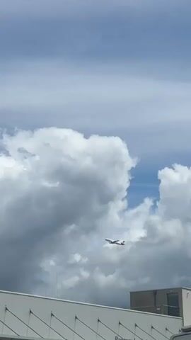 A passenger jet with red and white markings is seen flying against a sky filled with large, fluffy white clouds. Below the aircraft, the top of a white building with a modern design is visible. A white vehicle is parked in the foreground, partially obscuring the lower portion of the building. The camera angle is looking upwards towards the sky.