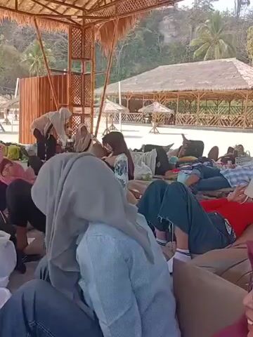 The video shows a group of people lying on beanbag chairs under a thatched-roof shelter on a sandy beach. Some people are looking at their phones, while others appear to be relaxing or sleeping. In the background, there are more thatched-roof structures and palm trees. The overall atmosphere appears to be casual and relaxed.
