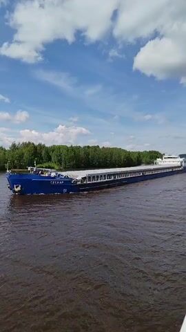 The video shows a large, dark blue barge moving from left to right across a wide, brown river. The barge has "СЕСКАР" written on its front side.  A row of green trees lines the riverbank on the left, while a white passenger boat follows behind the barge. Above, the sky is bright blue with scattered white clouds.  The water is slightly choppy, and the overall impression is of a bright, sunny day on a navigable waterway.