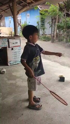 A young boy in a black t-shirt and beige shorts, holding a badminton racket, stands in a yard and swings it. He is wearing flip-flops. A black object is on the ground to his right. Behind him is a blue building with white trim and a thatched roof. To his left is a cardboard box. Another person is visible in the background.