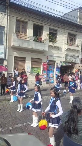 The video shows a group of young female dancers, dressed in matching navy blue and white uniforms with white boots, participating in a parade on a cobblestone street. They are carrying pom-poms in light blue and yellow. Buildings line the street with people watching from the sidewalks. A red car is parked at the front of the parade. One woman is taking pictures with her phone. The sky is overcast.