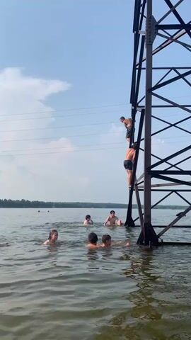 A group of young people are swimming and climbing on a large metal structure that appears to be a power line tower, partially submerged in a body of water. The sky is blue with scattered clouds. On the tower, one person is at the very top, and another is climbing the structure. Several other individuals are in the water, some swimming and others watching the climbers. The water is calm and appears to be a lake or river.