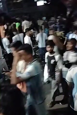 A crowd of people, many wearing blue and white striped clothing, are gathered in a public space at night. The video is shaky, suggesting it was filmed on a mobile device while the person was moving. Some people are holding or blowing into vuvuzelas, a type of horn. The atmosphere appears to be festive or celebratory. There are some lights and signs visible in the background, indicating an urban or commercial area. The scene is crowded and somewhat chaotic.