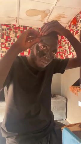 A young man with dark skin and a beard, wearing a black t-shirt, is in a room with patterned wallpaper. He is peeling a black face mask off his face, starting from his forehead and working down towards his nose and cheeks. He uses both hands to carefully remove the mask, focusing on the areas around his eyes and nose. The ceiling above him has several large brown stains.
