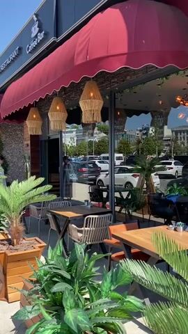 A sunny, high-angle outdoor shot focuses on the right side of a restaurant with outdoor seating. The left side features a red awning, and a sign for "Coffee Bar" can be seen. Below the awning are several decorative straw lamps. The restaurant has large glass windows that reflect a parking lot with cars and some buildings in the background. The interior of the restaurant is visible through the windows, showing tables, chairs, and tropical plants. Beyond the restaurant's interior, a body of water with distant ships can be seen. In the foreground, there are more tropical plants, and several tables and chairs for outdoor dining. The building's exterior is clad in light-colored wooden panels.