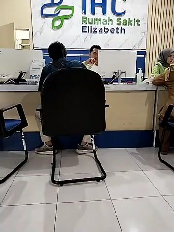 The video shows a scene inside a hospital registration area. A man wearing a dark shirt and light-colored pants sits in a black office chair facing away from the camera, towards a reception desk. He has a dark bag slung across his shoulder. Two other people are visible behind the reception desk, interacting with computer monitors. To the right, a woman wearing a light green headscarf and brown jacket is seated at the desk. The reception desk has white countertops, blue accents, and a large sign above that reads "IHC Rumah Sakit Elizabeth". The floor is tiled with large white squares. Chairs are visible in the foreground on either side of the man.