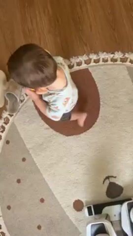 A toddler with short brown hair wearing pajamas stands on a round, cream-colored rug with brown accents and a fringed border, next to a pair of slippers. The toddler picks up a small, round yellow object, puts it in their mouth, and then walks away. The floor beneath the rug is wooden. Part of a white toy or object is visible in the lower right corner of the frame. The camera angle is positioned directly above the scene.