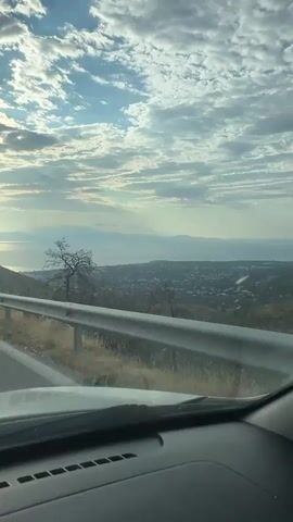 The video shows a view from inside a vehicle driving on a road. The road is paved and has white lane markings. A metal guardrail runs along the right side of the road. The landscape includes hills covered with dry vegetation and scattered trees. In the distance, there is a body of water, likely the sea, with islands or landmasses visible on the horizon. The sky is partly cloudy with the sun peeking through, creating a bright glow on the water. The overall scene suggests a scenic drive in a coastal or mountainous region during daytime, possibly early morning or late afternoon due to the lighting. No people or other vehicles are clearly visible.