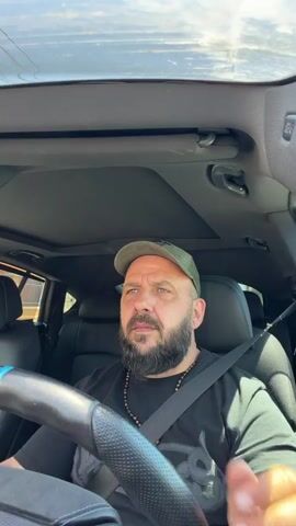 A mid-shot shows a man driving a vehicle. The man has a beard, is wearing a black t-shirt and a green baseball cap. He is wearing a seatbelt and a necklace. His left hand is on the steering wheel. The interior of the car is black, and there is a sunroof. The video is shot from the passenger seat.