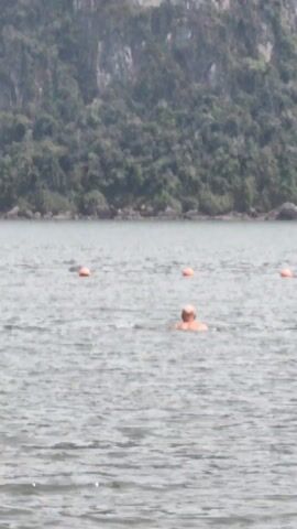 A person swims in a body of water, in the distance, with four orange buoys nearby. The swimmer appears to be wearing an orange top. Behind the water is a densely vegetated, steep rock formation. The day appears overcast.