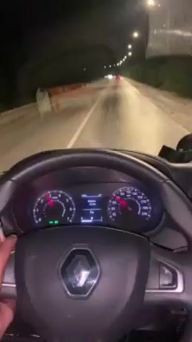 A first-person perspective video shows a driver holding a cigarette and steering a Renault car on a wet highway at night. The road is illuminated by streetlights, with other vehicles visible in the distance. The car's dashboard is visible, indicating a speed of around 100 km/h.