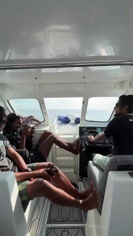 Inside a white cabin boat on the open sea, four men are visible. The driver, on the right, wears sunglasses and a black shirt with writing, focusing ahead. One man next to him has light green shorts. The second man from the left holds a camera and has his legs resting on the boat's structure. The man to his left is looking at his phone and the back of his head is visible. The sea and sky are visible through the boat's windshield.