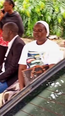 The video shows a group of people outdoors in what appears to be a casual setting, possibly near a building with lush green foliage. In the foreground, a woman in a white t-shirt with a colorful graphic print is seated, looking directly at the camera. She is wearing a white head covering. Beside her, a man in a dark suit jacket and blue jeans is also seated, looking to his left. Further back, a woman in a black headscarf is standing and talking on a mobile phone. The backdrop features dense green leaves and some light-colored walls, suggesting a warm climate. The overall mood seems relaxed, and the lighting indicates it is daytime. There are no visible signs of a specific event beyond people gathered, though the presence of a car in the foreground hints at transportation or waiting.
