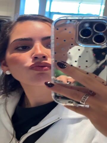 A young woman with dark hair and fair skin is featured in a mirror selfie video. She wears a white zippered jacket with a Ralph Lauren polo emblem over a black top and a pearl earring in her left ear. Her nails are painted a dark shade, possibly deep red or black, as she holds an iPhone in a clear case with colorful polka dots. She shifts her facial expression from a slight smile to a playful pout. A ring is visible on her right hand, and a red band is partially visible on her left wrist. The background shows blurred mannequins and people, indicating an indoor retail store setting in San Marcos, United States.