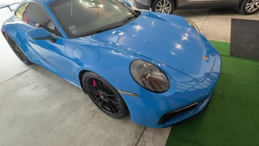 The video begins with a high-angle shot of a bright blue Porsche 911 sports car. The camera slowly pans across the front and side of the car, focusing on the sleek design, black wheels, red brake calipers, and the Porsche emblem on the hood. The car is parked indoors, likely in a garage or showroom, with a concrete floor and a green carpet visible on the right side of the frame. In the background, another car, partially visible, appears to be a grey SUV. The lighting is bright, reflecting off the car's glossy surface. There are no people visible in the video, and no actions other than the camera movement. The timestamp on the screen indicates the video is just a few seconds long.