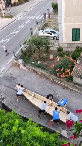 Six men are carrying a large wooden canoe, likely a homemade one, down a sloped asphalt driveway. The canoe is long and narrow, made of unfinished plywood. The men are positioned along the sides of the canoe, with two at the front, two in the middle, and two at the back, all lifting it together. They are all wearing casual athletic clothing, including t-shirts and shorts. One man, possibly a supervisor or director, stands on the left side of the driveway, observing the group. The driveway is bordered by a low retaining wall on the left, with green foliage and red flowers in the foreground. On the right, there's a building with a green shuttered window and a small garden area with flowering bushes and trees. Across the driveway is a street with a parked silver car and a sidewalk with a white fence. The scene appears to be a residential area, possibly in Italy given the architecture. The men are moving slowly and carefully, coordinating their steps to carry the heavy object.