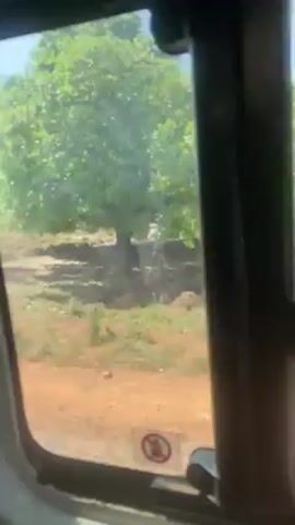 The video shows the view from a car window. The car is moving along a dirt road, passing by fields and buildings. The buildings appear to be houses, and there are trees visible. The sky is clear. A no smoking sign is visible on the window.
