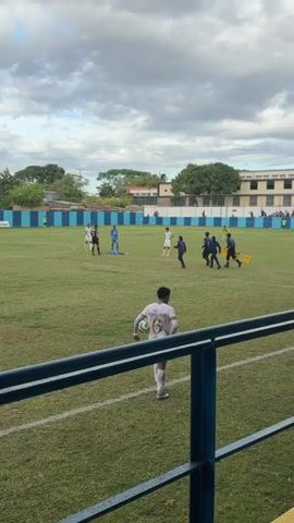 A video shows a football match interrupted by an injured player on the ground, surrounded by medical personnel and other players. The player is dressed in a light blue jersey and is lying on the green grass. Medical staff are attending to the injured player, with some carrying a stretcher. Other players stand nearby, observing the situation. The background features a blue and white striped wall, buildings, and trees under a cloudy sky. One player in a white jersey and number 6 is visible in the foreground, looking towards the scene.