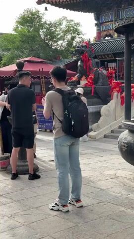 The video shows a courtyard scene outside a Chinese temple. A man wearing a tan shirt, light blue jeans, and a black backpack lights incense sticks and places them in a large incense burner. There are other people in the background, including a man in a black shirt and shorts who appears to be waiting to light incense. The temple is decorated with red ribbons, and a dragon statue is also visible. Smoke rises from the burner.