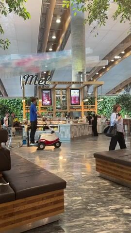 The video shows an indoor scene, likely an airport or large public space, with a bakery counter called "Mel Norwegian Bakery". Several people are visible. A person in a blue shirt is standing near a small, red and white vehicle. Another person in black is behind the counter, possibly working. A person with dark hair and a light-colored shirt is visible on the right side of the frame, and another person with blonde hair is on the left. Rows of luggage carts are lined up in front of the bakery. The environment features large columns, bright lighting, and decorative greenery hanging from the ceiling. The floor is a polished stone or marble.