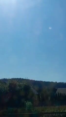 The video shows a view from inside a vehicle moving through a residential area on a sunny day. The sky is clear blue with some light flares from the sun. Trees and houses are visible on either side of the road. The houses appear to be made of concrete or stucco, with tiled roofs and various window styles. Some houses have balconies, and there are fences and some greenery. The vehicle appears to be moving at a moderate speed. The audio is in Russian and states "In 300 meters, at the roundabout."