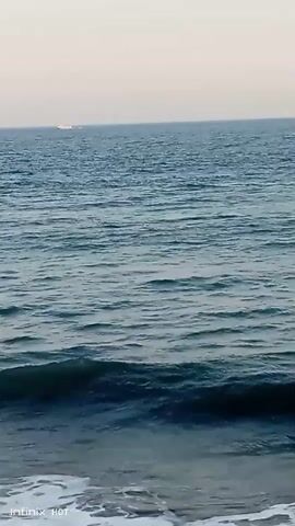 The video shows a view of the ocean from a shoreline. The water is blue and appears to have moderate waves. The sky is a pale, hazy color, suggesting it might be dawn or dusk. In the distance, a white object, possibly a boat or a distant structure, is visible on the horizon. The foreground shows the white foam of waves breaking on the shore. There are no people or specific objects clearly identifiable in the scene apart from the natural elements of the sea and sky. The video appears to be a short, static shot of the ocean.