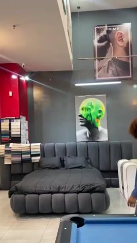 A person with curly hair walks across the screen towards a grey tufted bed and a pool table. The room appears to be a modern showroom or salon with various artistic posters on the walls, including a poster of a man's ear being cut, a distorted face with green and yellow colors, and a woman's face. There is also a bright red wall with geometric patterns and a pool table with billiard balls and cues in the foreground. The lighting is bright and overhead.