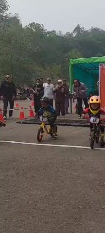 Two young children, wearing helmets, are participating in a race on balance bikes. One child, on the left, wears a yellow and blue jersey with the number 21 on their bike. The other child, on the right, wears a red and black jersey with the number 18 on their bike. They are on an asphalt road, with orange cones marking the racecourse. Spectators are visible in the background, some holding umbrellas. The environment appears to be outdoors, with trees and hills in the distance. The sky is overcast.