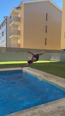 A person with dark hair wearing dark shorts is swimming in a rectangular blue pool. The pool has a light-colored tile border. The person is in the middle of the pool, with their head submerged and arms extended forward. Water splashes around them as they swim. Behind the pool is a patch of green grass, a gray wall, and two multi-story buildings with cream-colored and tan facades. The sky is clear and blue.